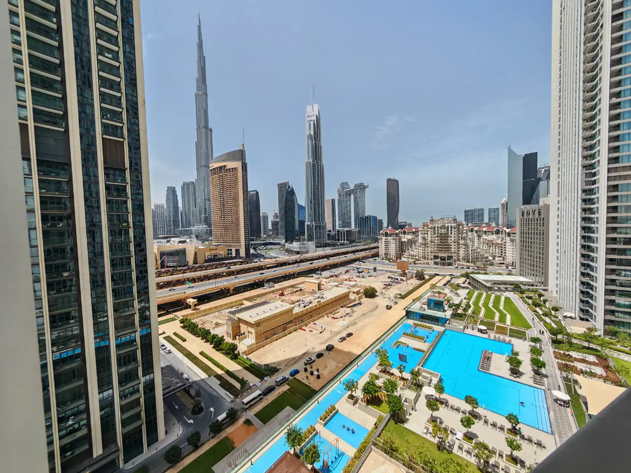 View of Dubai skyline featuring Burj Khalifa, skyscrapers, pools, and greenery from a high-rise balcony on a sunny day.