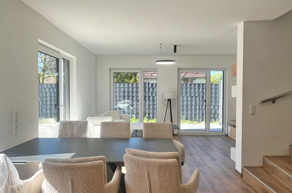 Bright, modern dining area with a gray table and beige chairs. Large windows offer a view of the backyard. Wood floors and stairs add warmth.