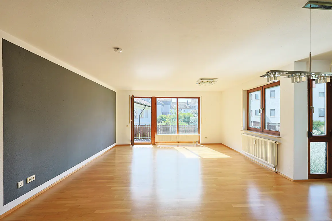 Bright, empty living room with wood floors, gray accent wall, and balcony access. Natural light floods the space.