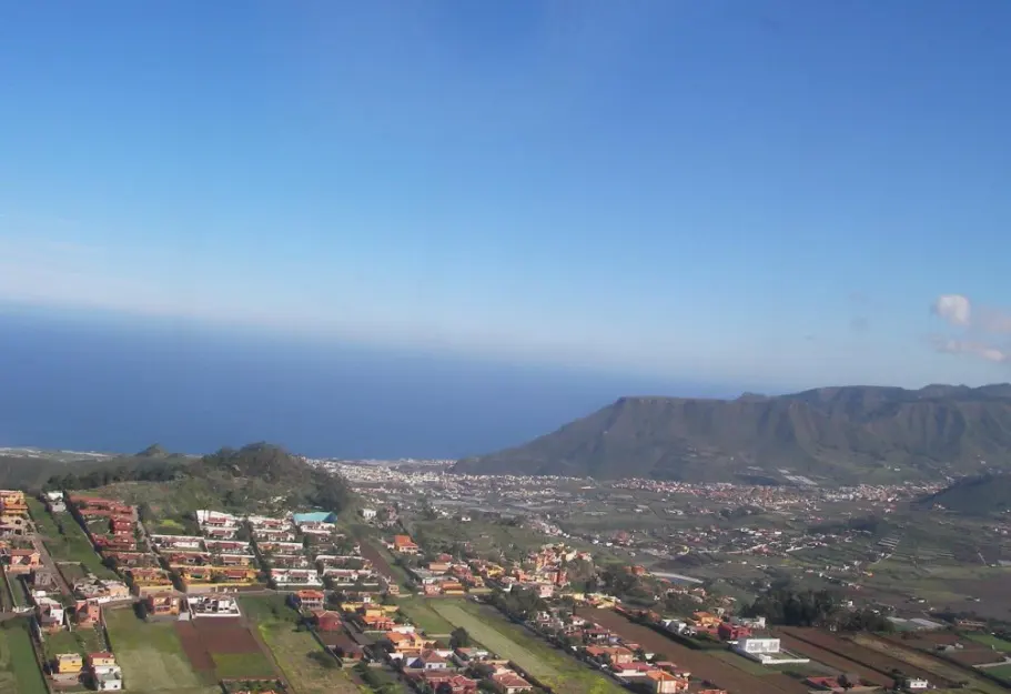 Aerial view of a coastal town with red-roofed houses, green fields, and mountains against a blue sky and ocean.