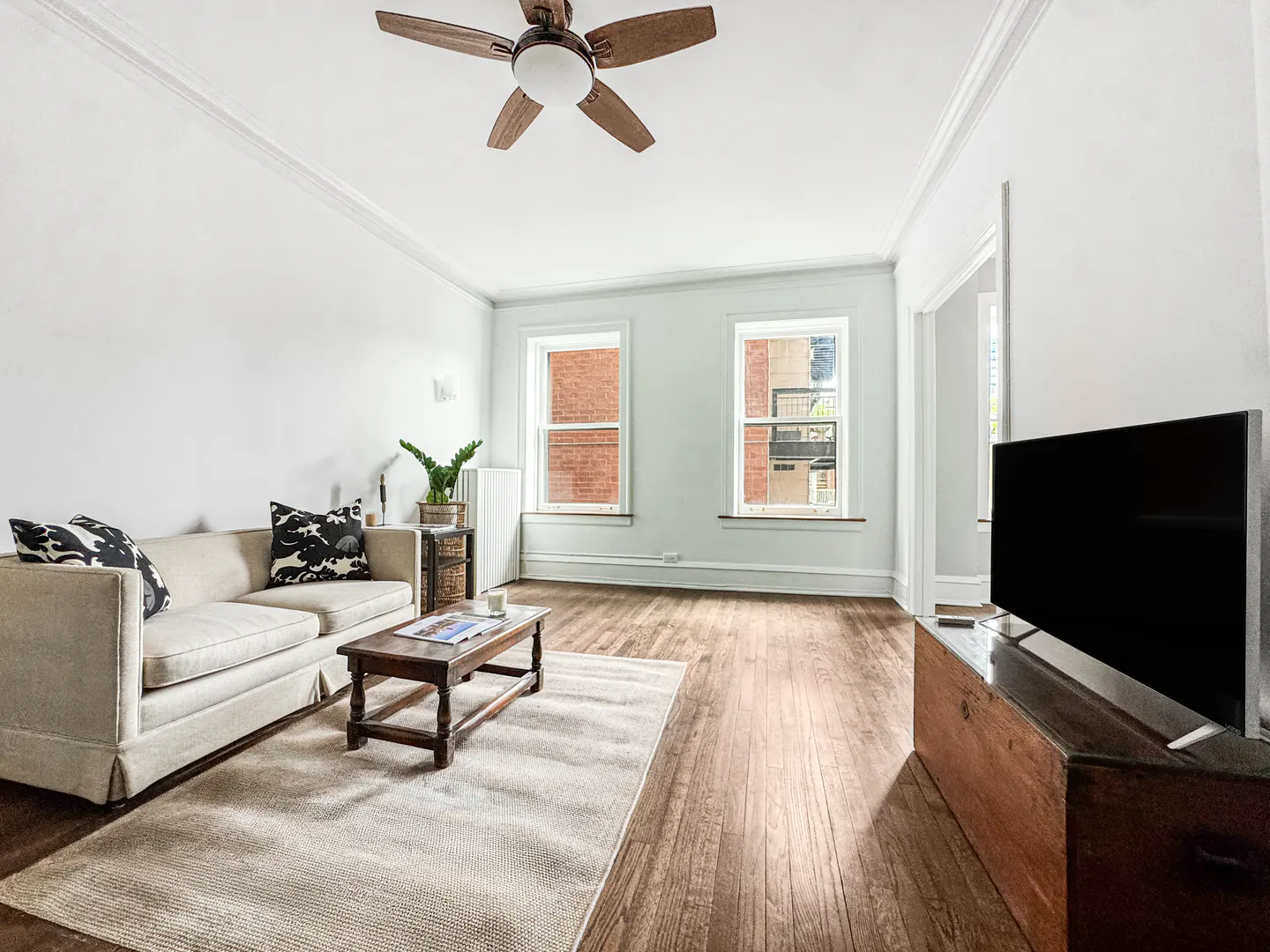 Living room with white walls, wood floors, and a ceiling fan. A beige sofa, wood coffee table, and TV stand are present. Two windows offer natural light.