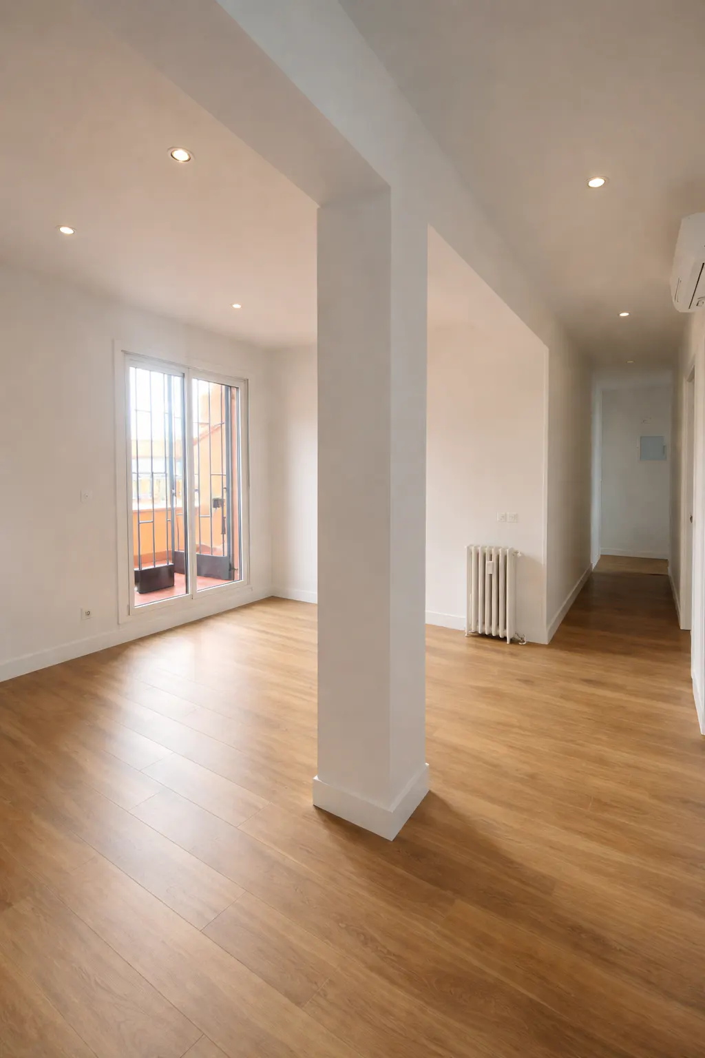 Bright, empty room with wood floors, white walls, and a large support column. Sliding glass doors lead to a balcony. A radiator is visible.