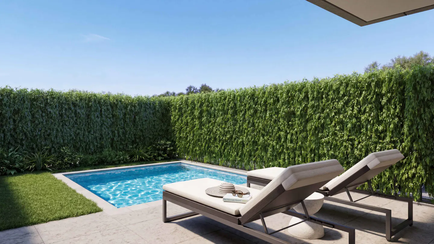 A backyard with a pool, two lounge chairs, and a tall green hedge. A hat, book, and sunglasses sit on one chair.