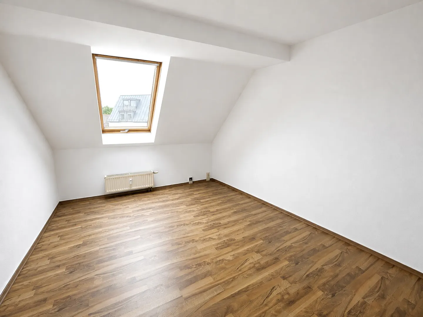 Bright, empty attic room with white walls, wood floor, and skylight. A radiator sits under the window.