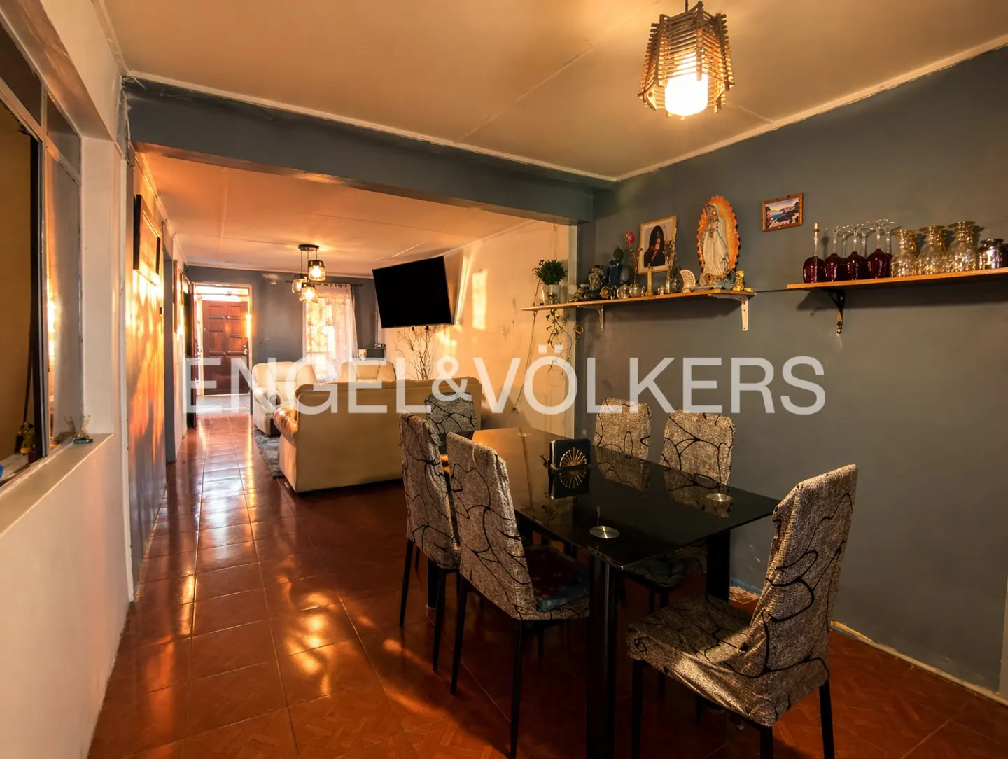 Interior view of a dining area with a black table and patterned chairs, leading to a living room with a white sofa. Shelves with decor on the wall.