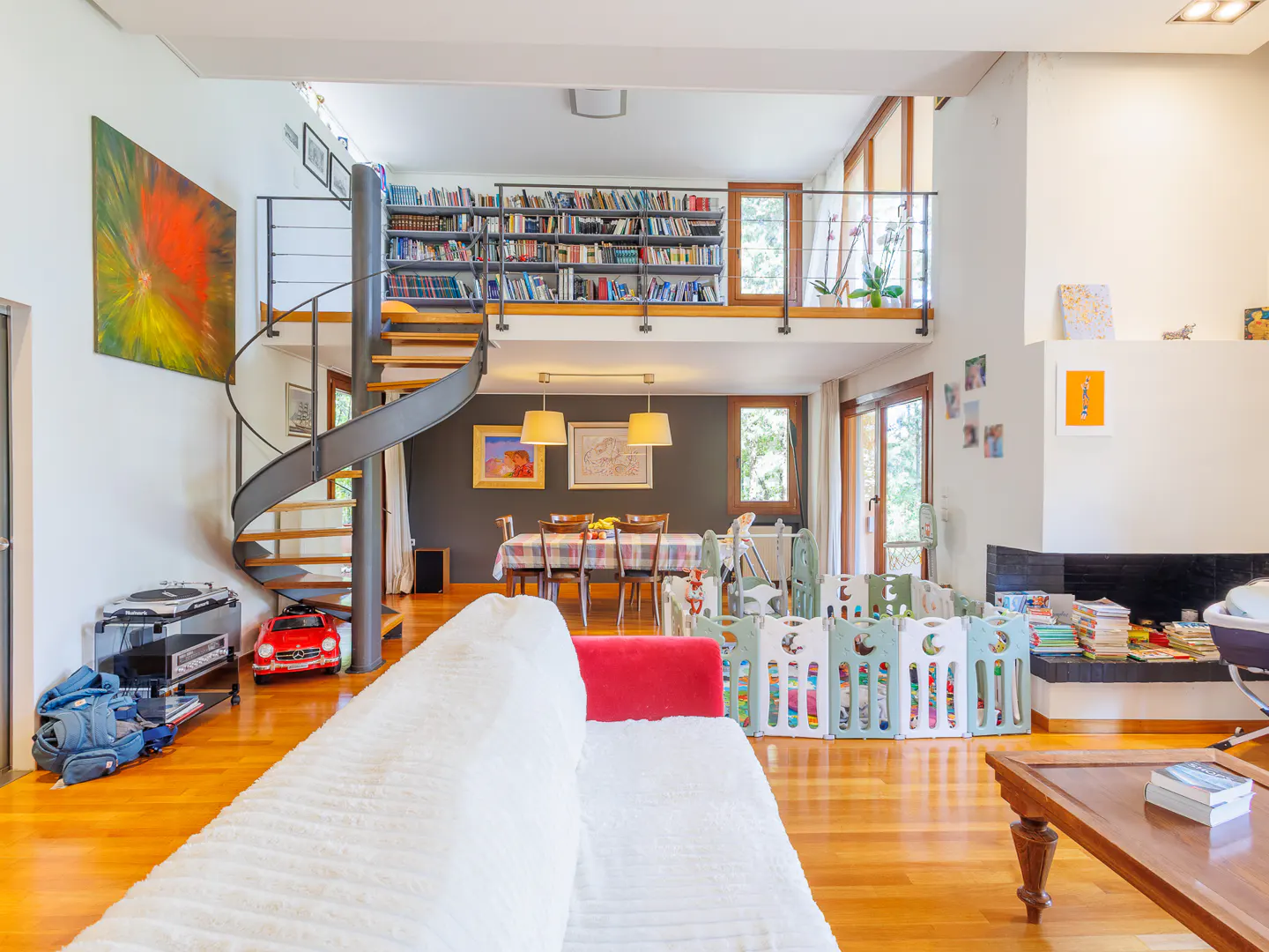 Bright living room with hardwood floors, a spiral staircase, and a loft with bookshelves. A dining table and a playpen are also visible.