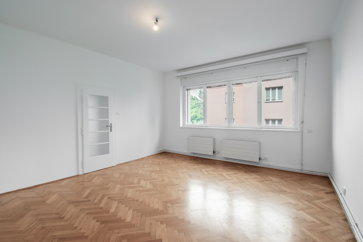 Empty room with white walls, herringbone wood floors, a white door with glass panels, and a large window with radiators.