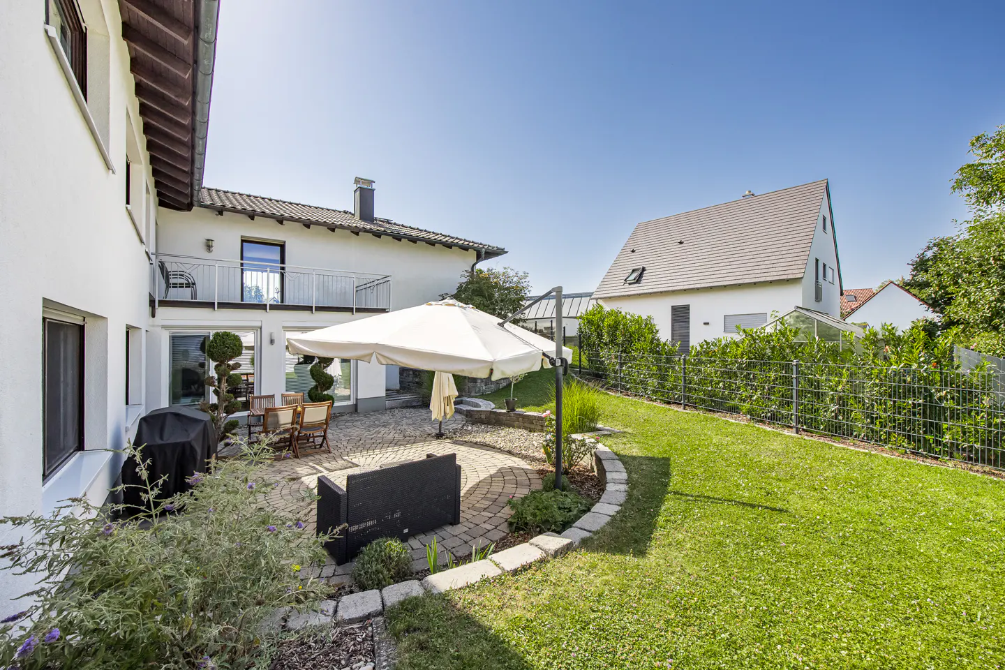 A backyard with a patio, lawn, and white houses under a blue sky. A large white umbrella shades a table and chairs.