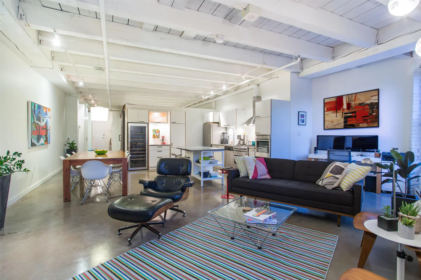 Open-concept loft with white wood-beamed ceilings, concrete floors, and a black sofa. A dining area, kitchen, and office space are also visible.