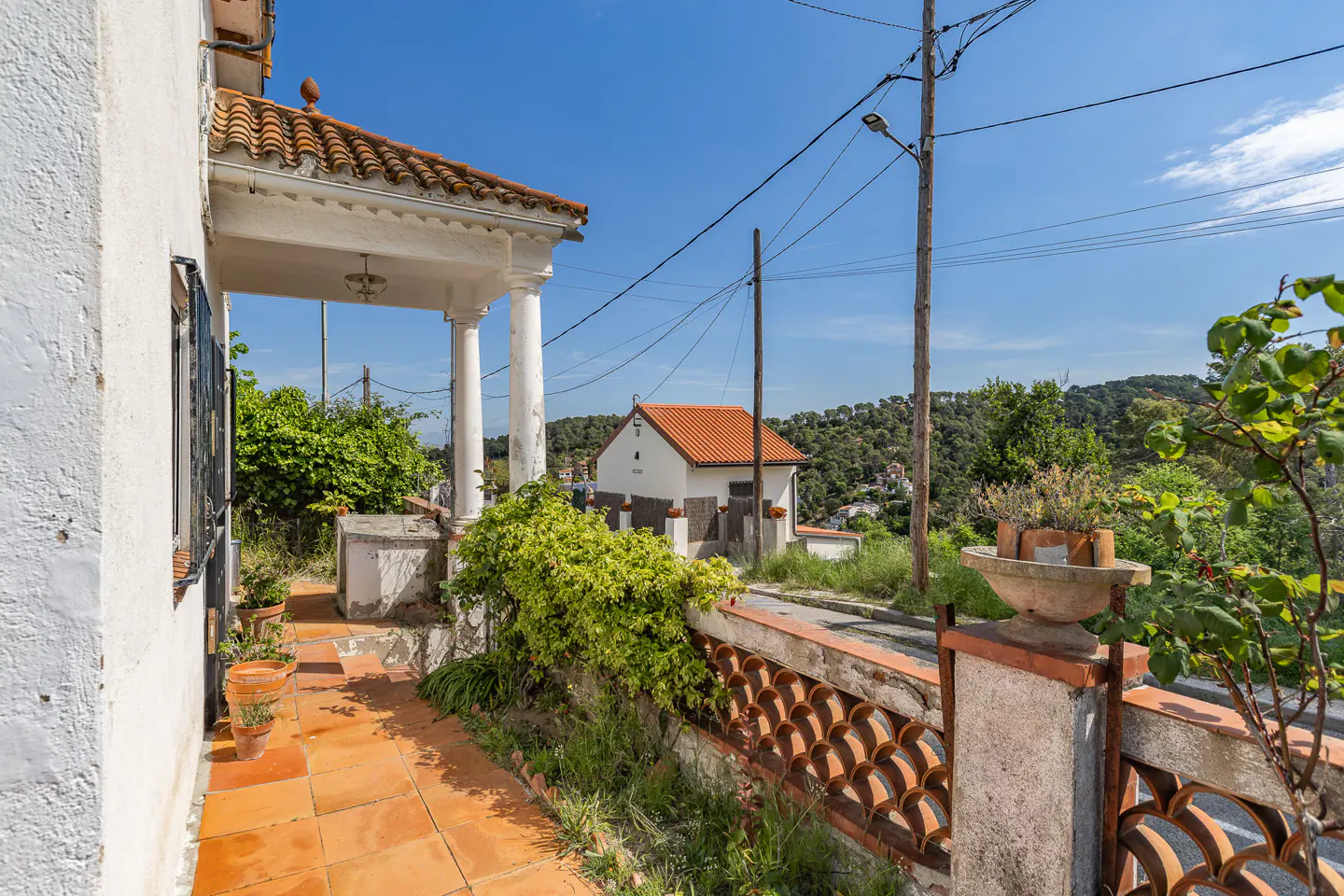 Exterior view of a white house with a tiled porch and decorative brick railing, set against a blue sky and green hillside.