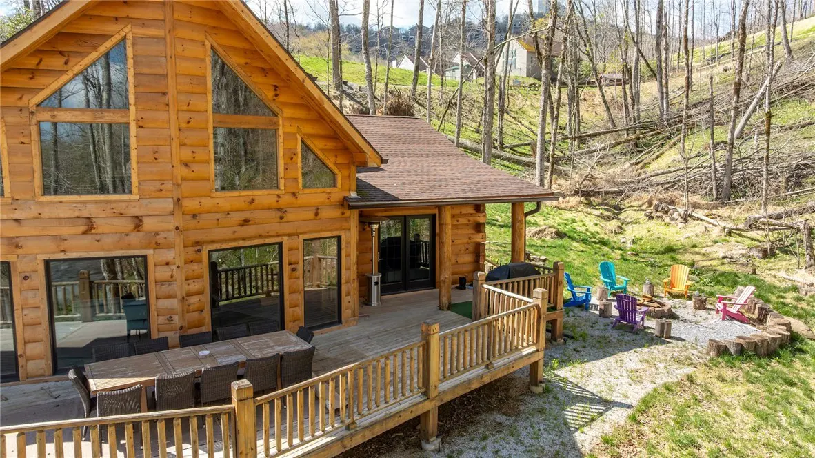 A rustic log cabin with a wooden deck, outdoor dining set, and colorful chairs in a wooded, hilly backyard.