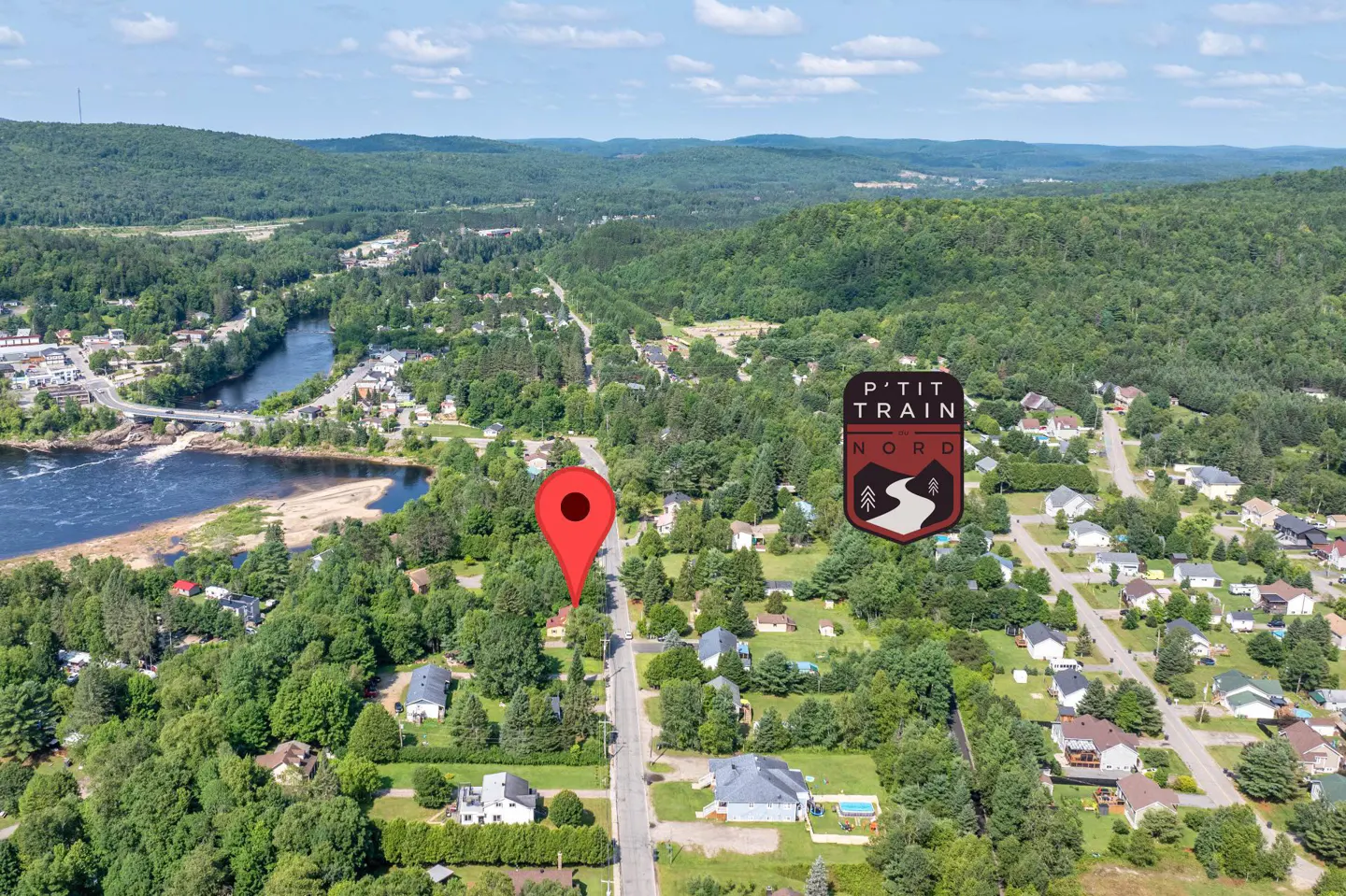 Aerial view of a residential area near a river and forest, with a red map pin marking a property near P'tit Train du Nord.