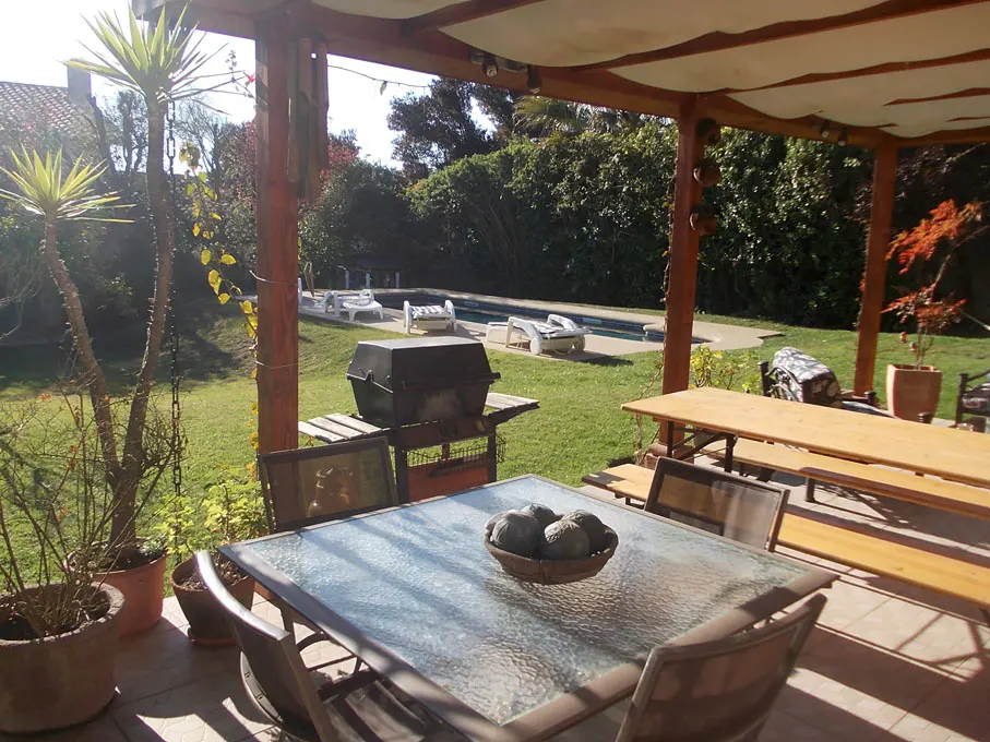 Outdoor patio with glass table, chairs, grill, and wooden picnic table. Pool and green lawn in the background.