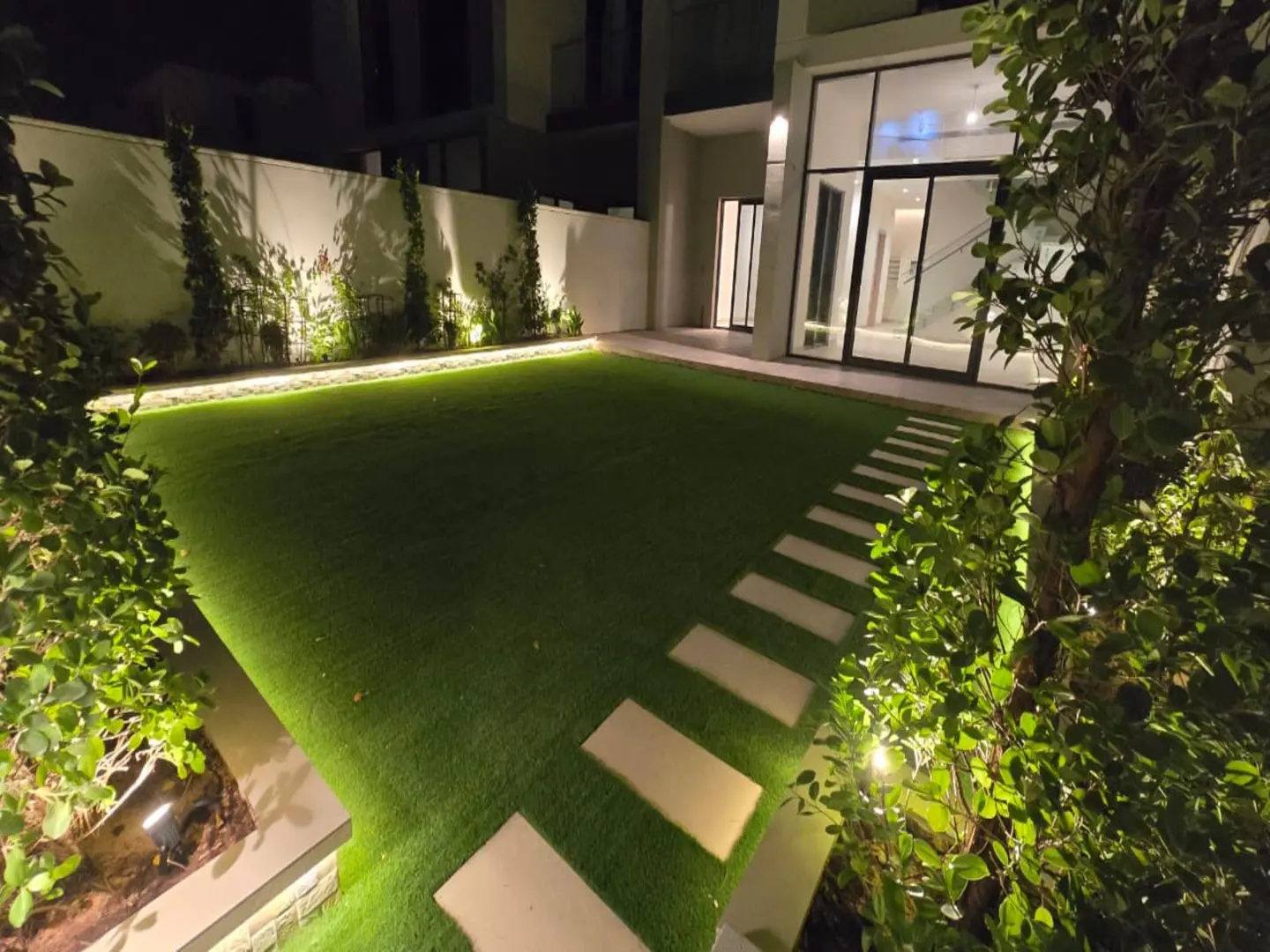Night view of a modern home backyard with a green lawn, stone stepping stones, and warm landscape lighting.
