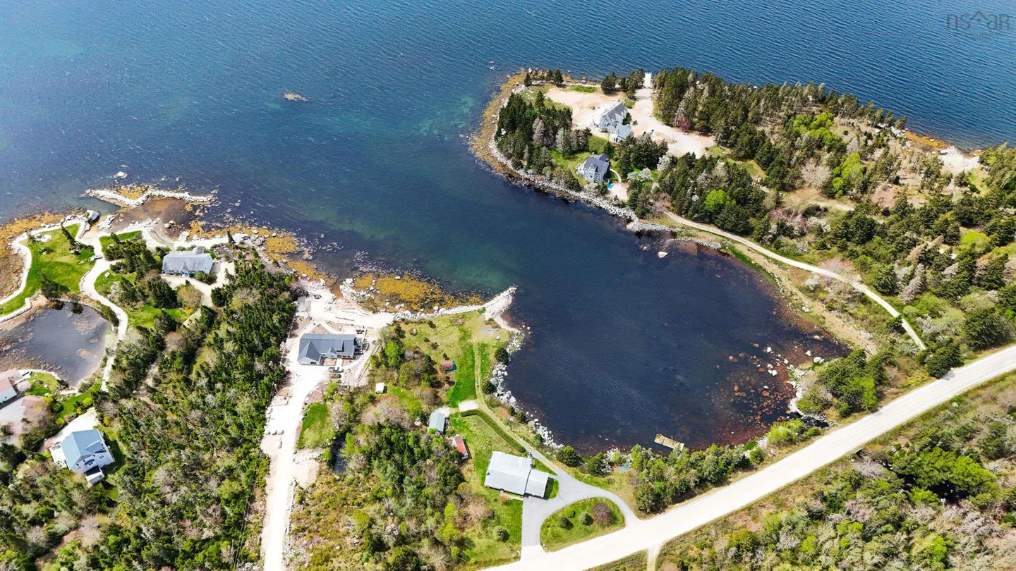 Aerial view of coastal real estate with houses nestled among trees and a calm bay. Blue ocean water surrounds the property.
