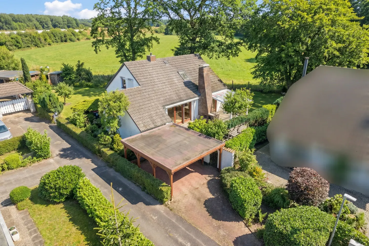 Aerial view of a white house with a brown tile roof, carport, and green lawn. Trees and a field are in the background.