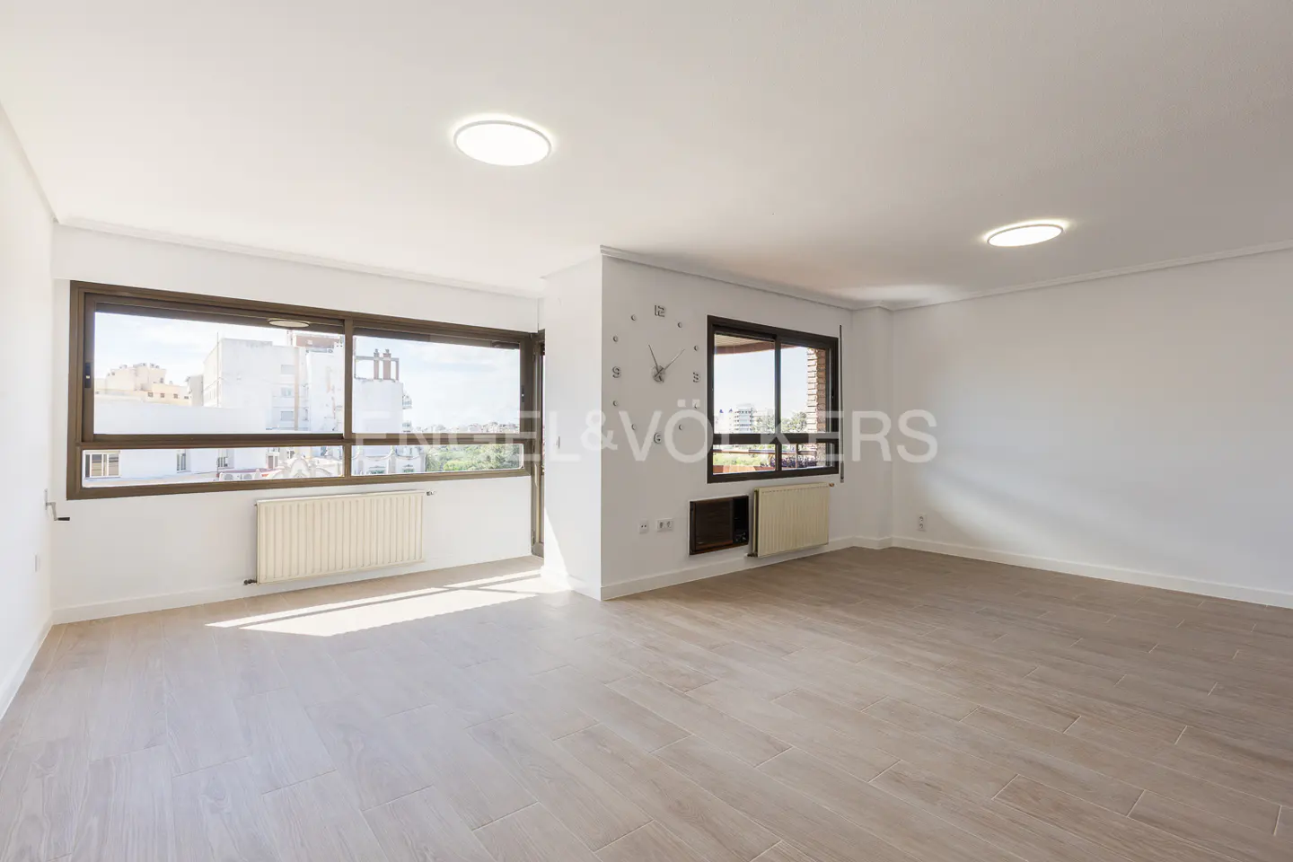 Bright, empty room with light wood floors, white walls, and two windows with brown frames. A clock is on the wall between the windows.