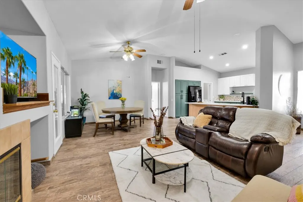 Open-concept living space with a brown leather sofa, white rug, and wood-look floors. A dining table and kitchen are in the background.