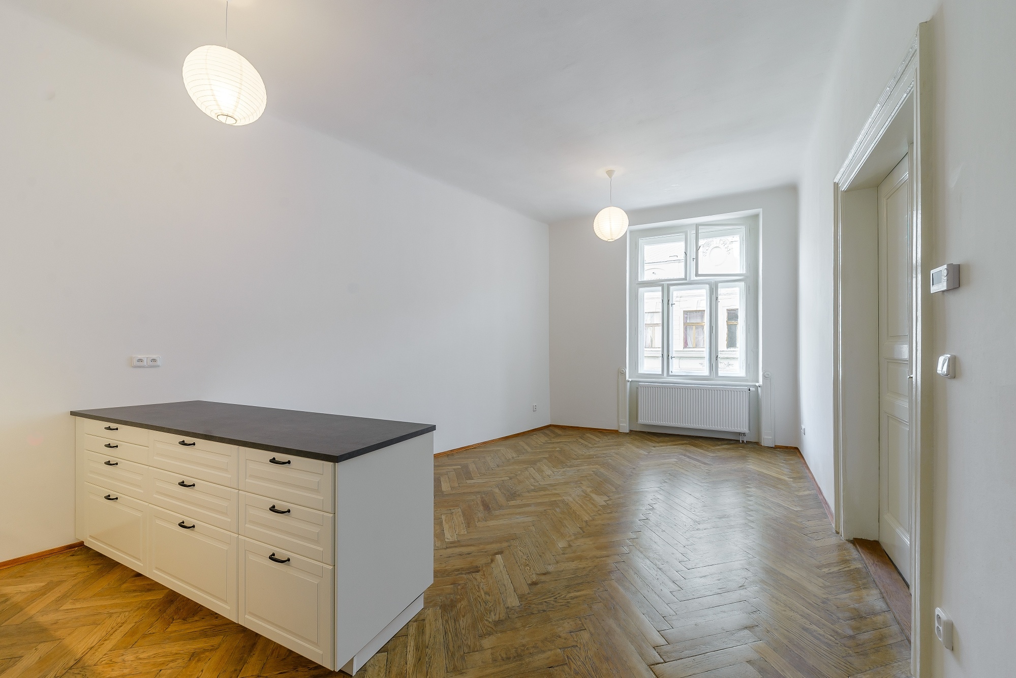 Empty room with white walls, herringbone wood floors, a white kitchen island with a dark top, and two globe pendant lights.