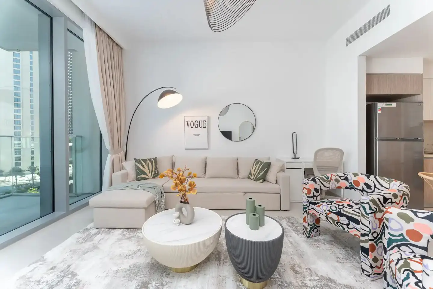 Bright living room with beige sofa, patterned chairs, and white walls. Large window with city view. Modern decor with round tables and Vogue print.
