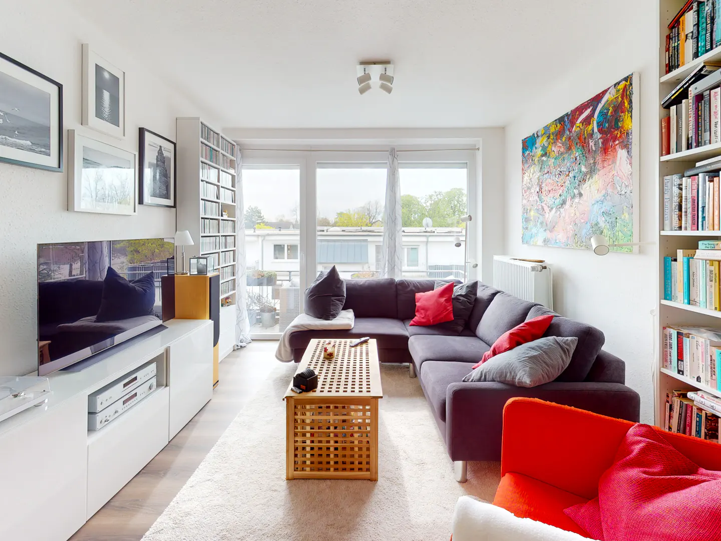 Bright living room with a gray sectional sofa, a wooden coffee table, and a white media console with a TV. Bookshelves and colorful art add character.