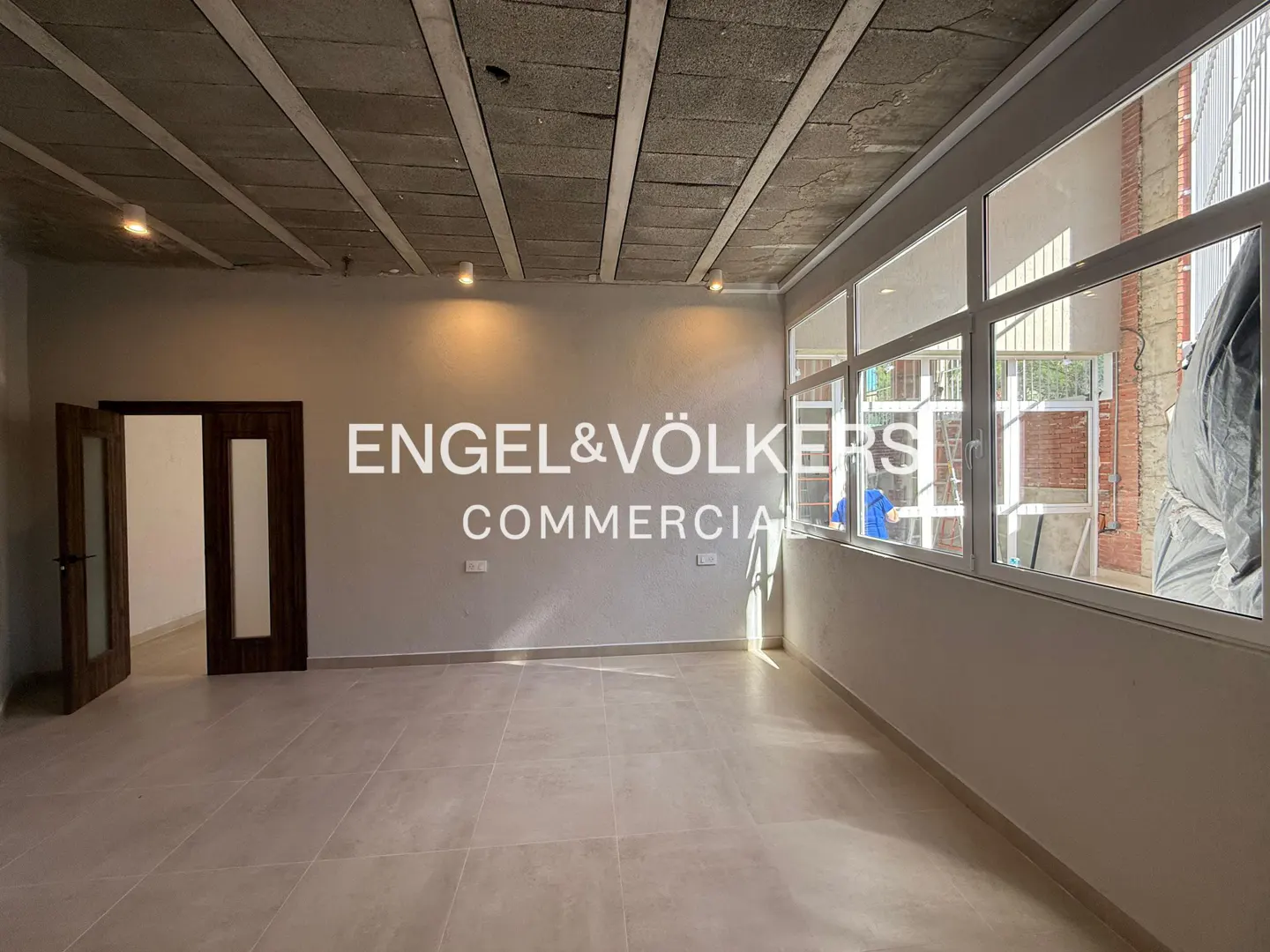 Empty commercial space with gray walls, tile floor, and exposed concrete ceiling. Large windows line one wall, and a dark wood door is on the left. "Engel & Völkers Commercial" logo is centered.