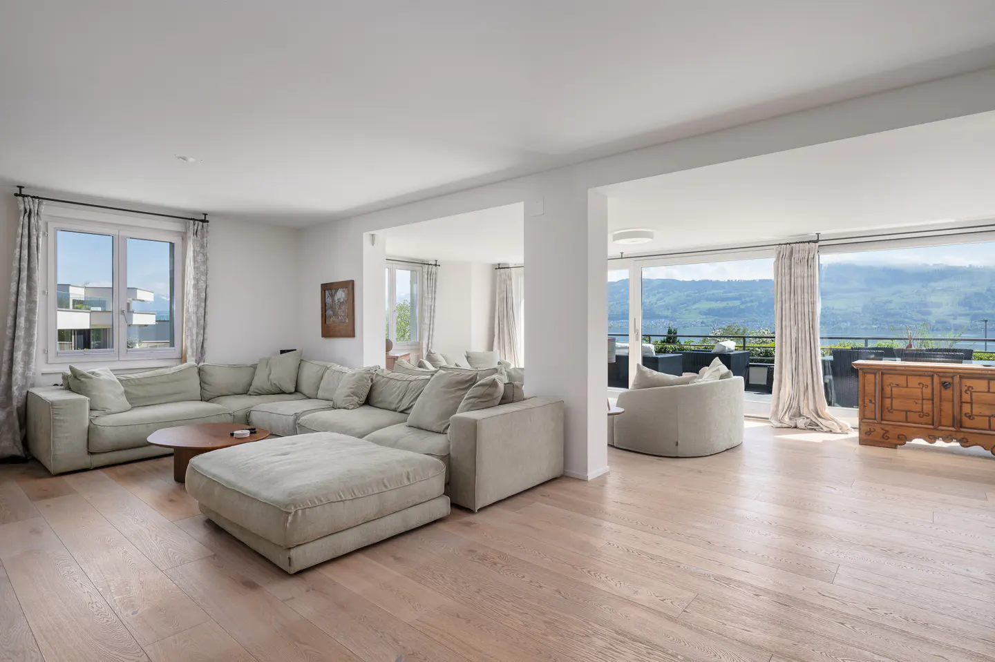 Bright living room with a large beige sectional sofa, wood floors, and a view of a lake and mountains through large windows.