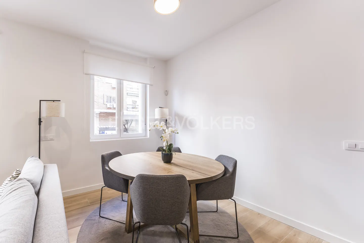 A bright dining area with a round wooden table, four grey chairs, a white orchid centerpiece, and a floor lamp.