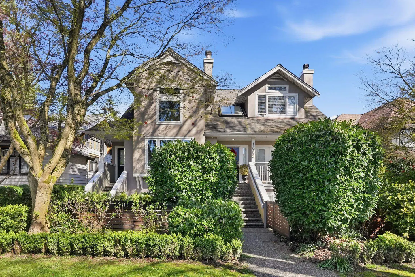 Two-story beige house with white trim, chimney, and landscaped yard on a sunny day.