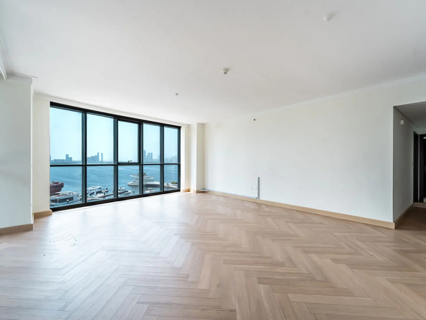 Bright, empty room with herringbone wood floors and white walls. Large window shows a harbor with boats and city skyline.