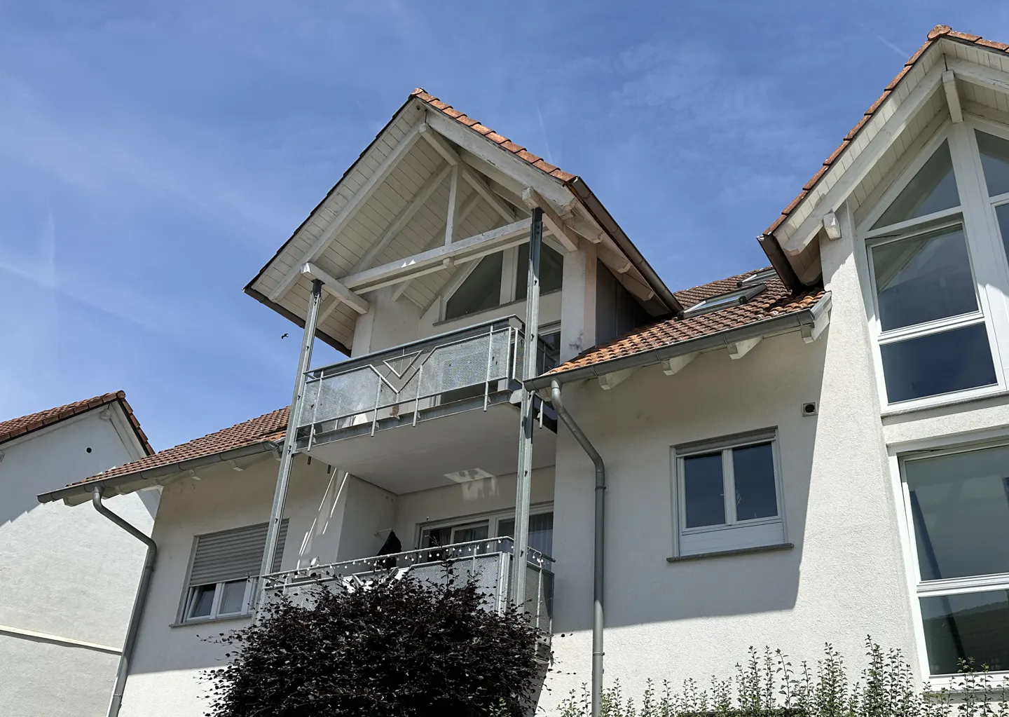 White multi-story residential building with balconies, gabled roof, and large windows under a clear blue sky.