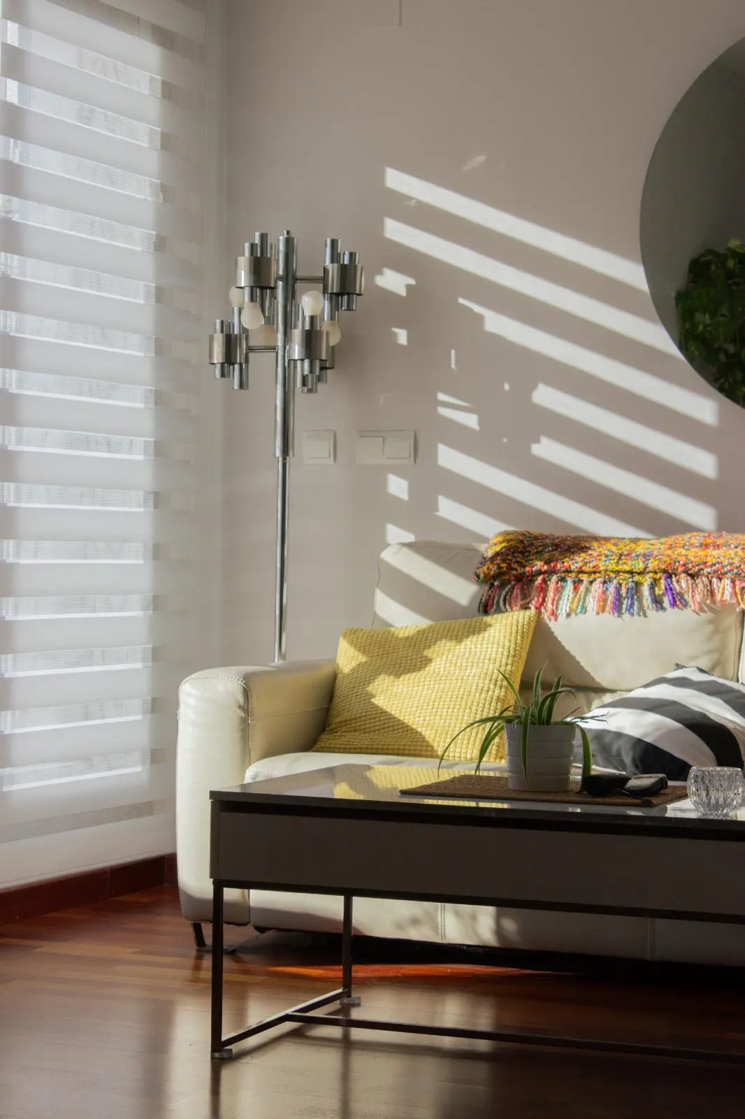 Sunlit living room with a white sofa, yellow pillow, and striped blanket. A modern lamp stands near a window with blinds.