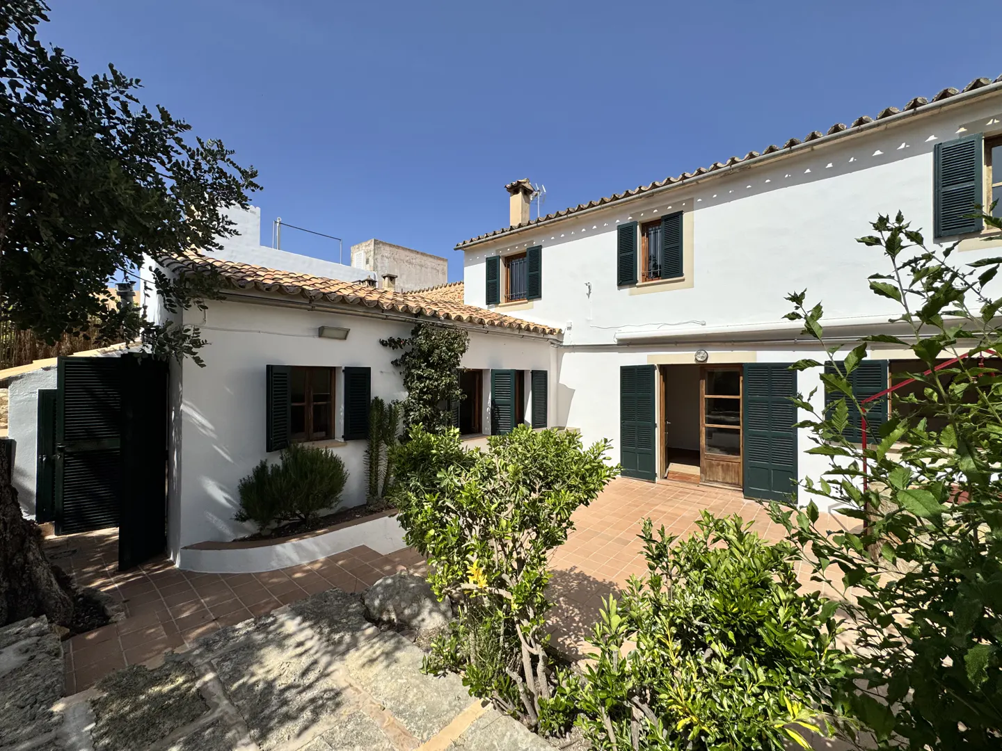 Exterior view of a white house with green shutters, a terracotta tiled roof and patio, and lush green landscaping.