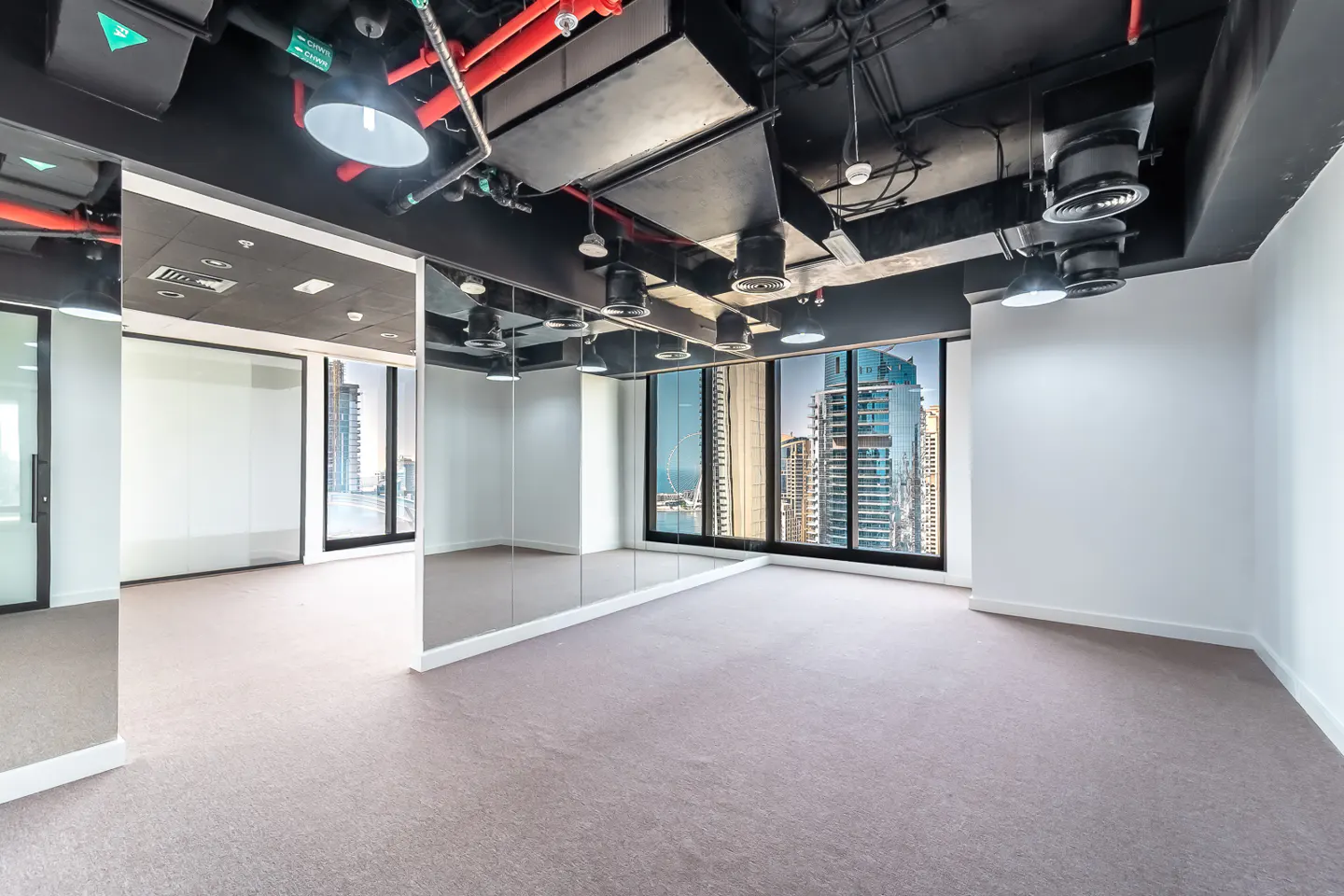 Empty office space with gray carpet, white walls, and a black ceiling with exposed pipes and ductwork. Large windows offer a city view.