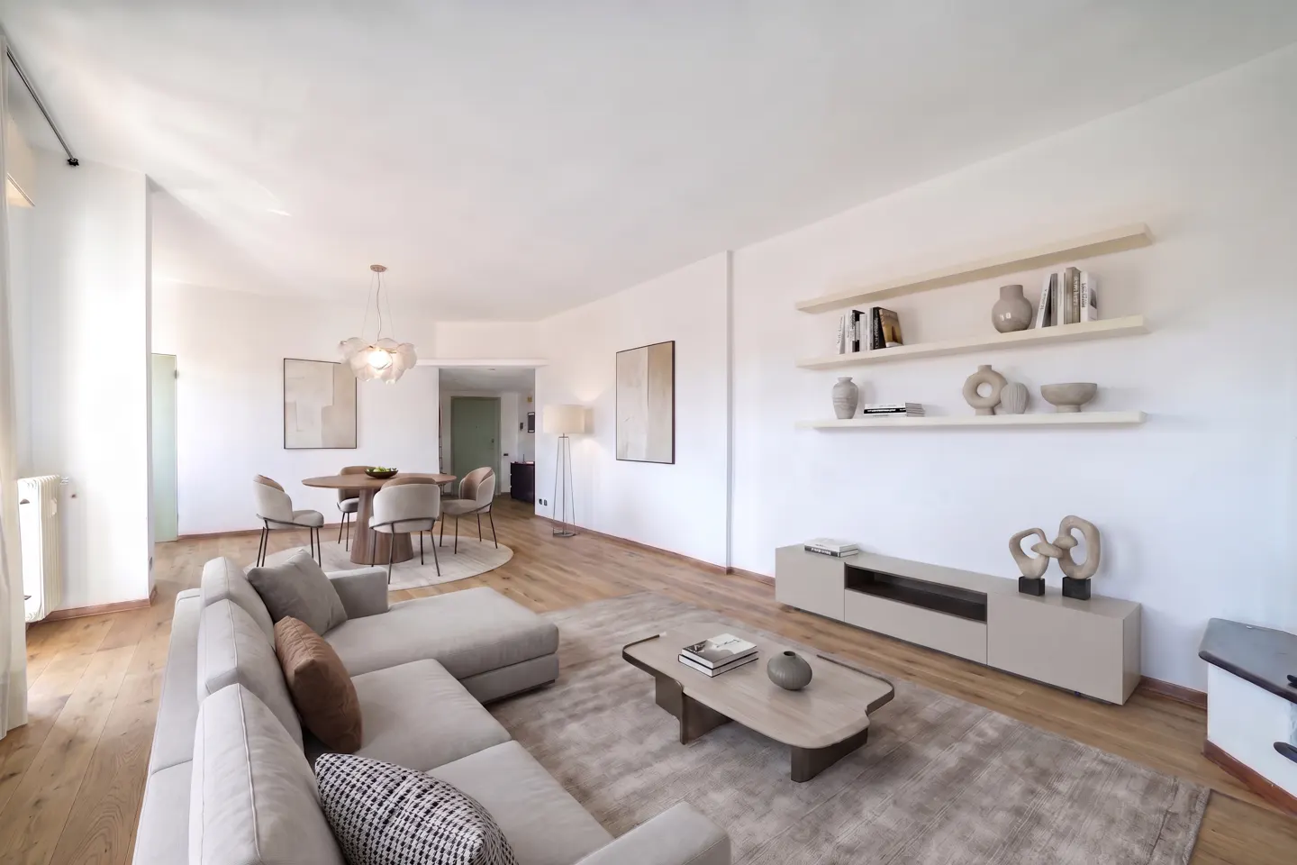 Bright, modern living room with a beige sectional sofa, wood floors, and white walls. A round dining table and shelves with decor add to the stylish space.