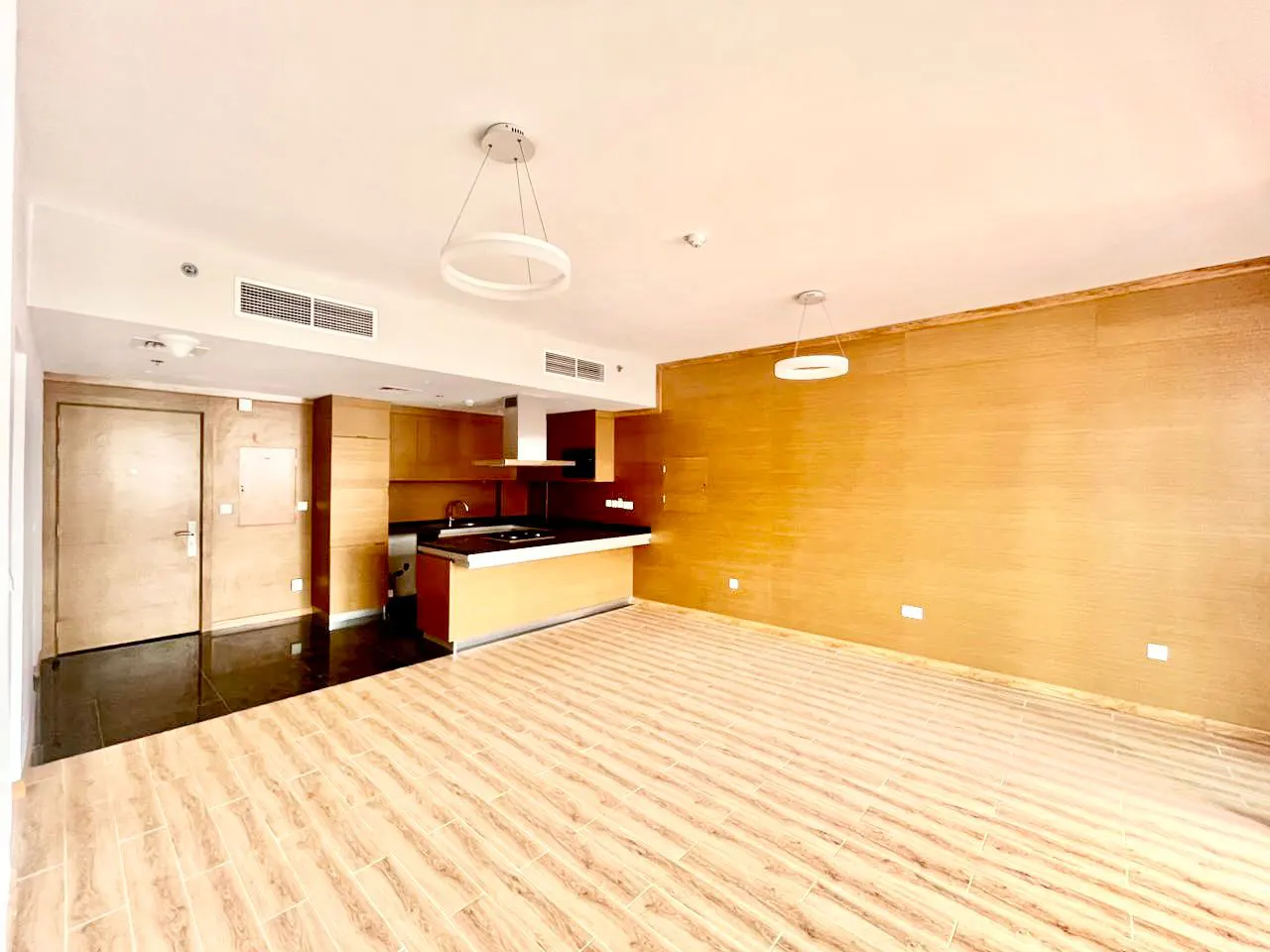 An open-concept apartment with wood floors, a kitchen island, and a gold accent wall. Two modern, circular pendant lights hang from the white ceiling.
