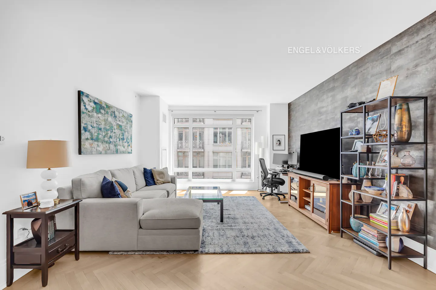 A bright living room with a gray sectional sofa, a large window, and a TV with a bookshelf.