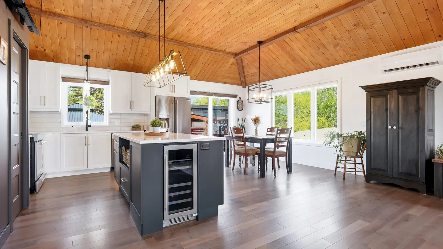 A bright, open kitchen and dining area with wood floors and a vaulted wood ceiling. White cabinets and a gray island with a wine fridge.