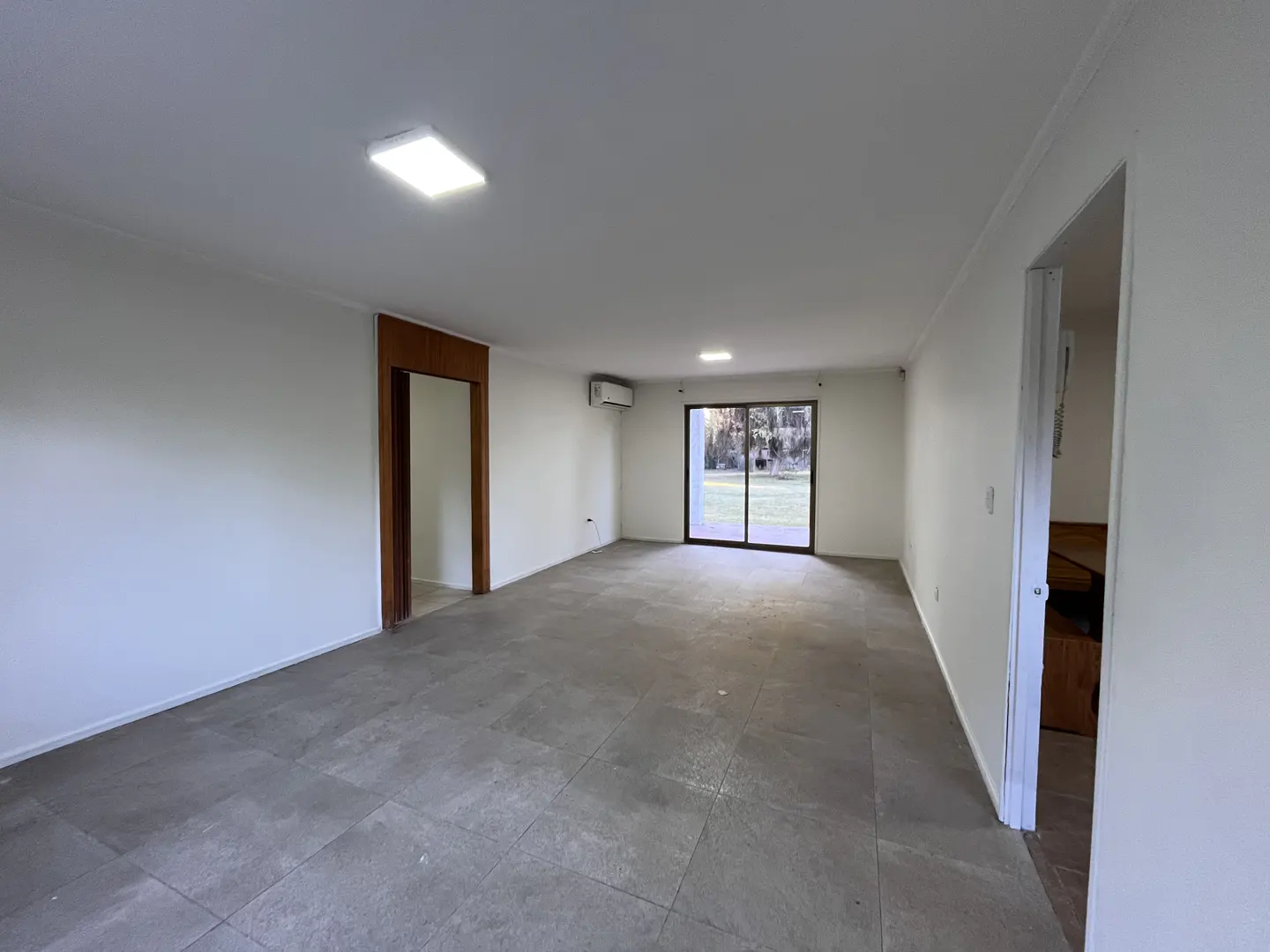 An empty room with white walls, gray tile floor, and a sliding glass door to the outside. A wooden door frame is on the left.