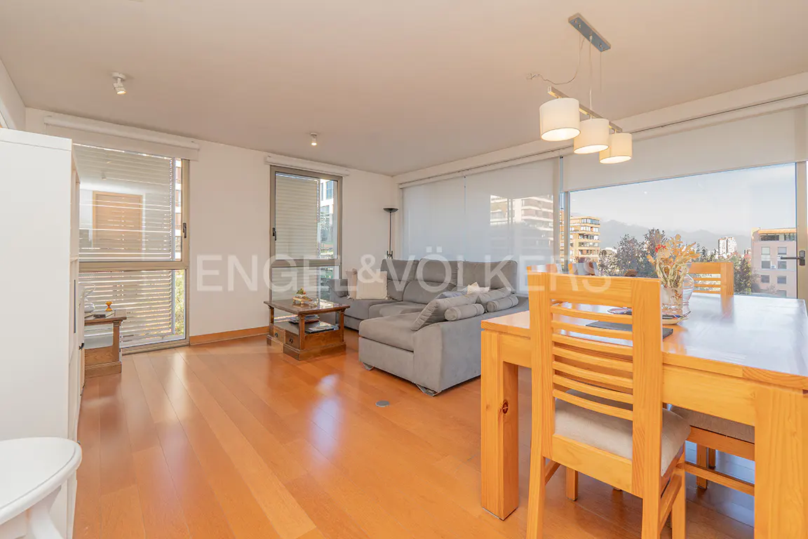 Bright living room with wood floors, gray sofa, and wood dining table with chairs. Large windows offer city views.