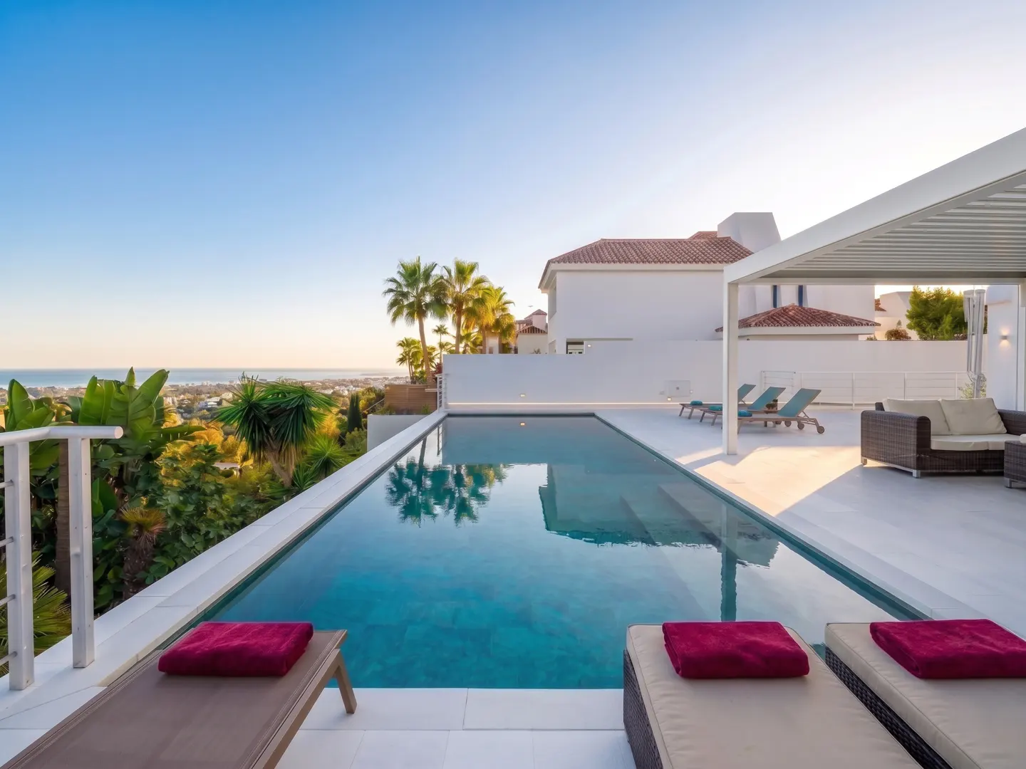 Infinity pool with lounge chairs and red towels overlooks a lush landscape and ocean under a clear blue sky. White modern house in background.