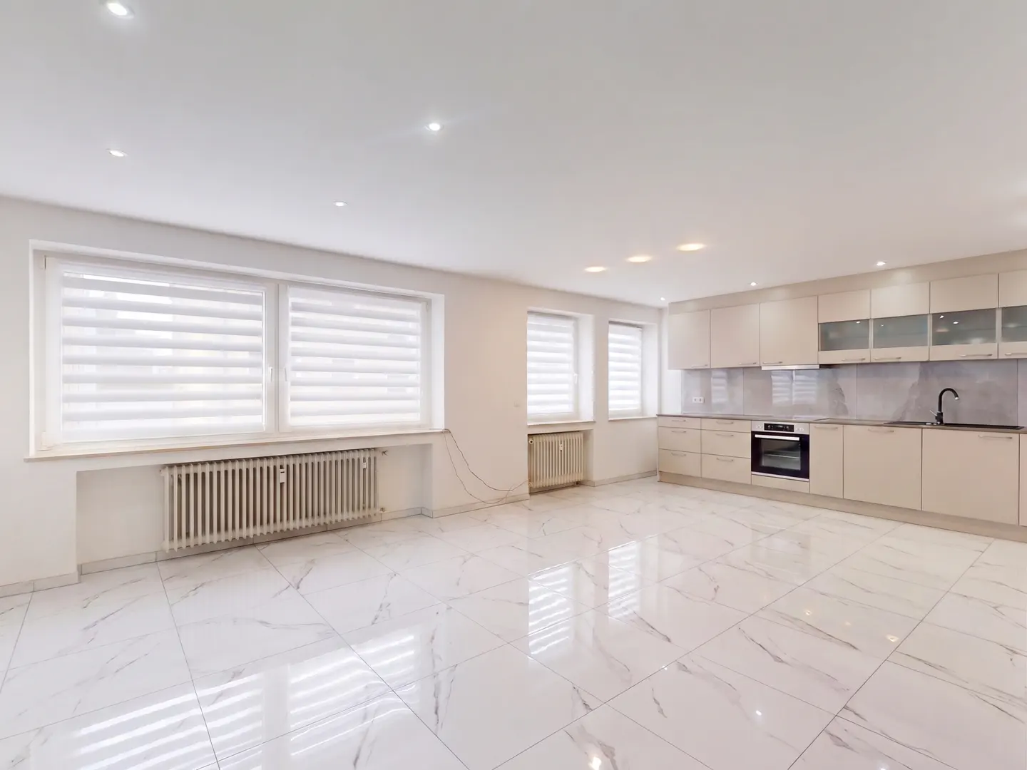 Bright, empty room with white marble floors, large windows with blinds, and a modern kitchen with beige cabinets.