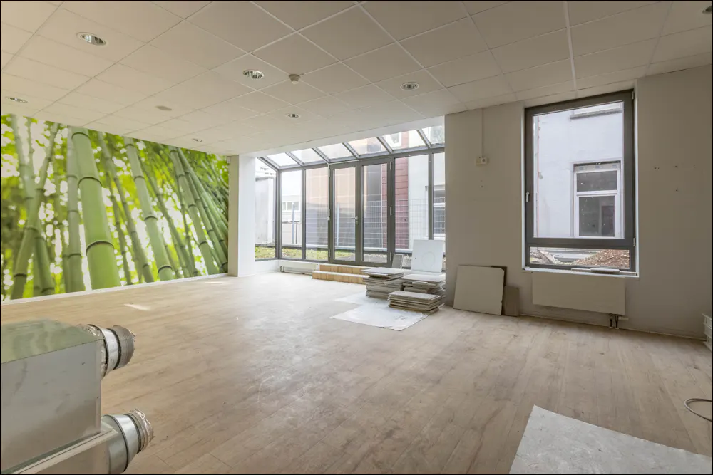 Bright, empty room with light wood floors, bamboo mural, and large windows. Tiles are stacked in the center of the room.