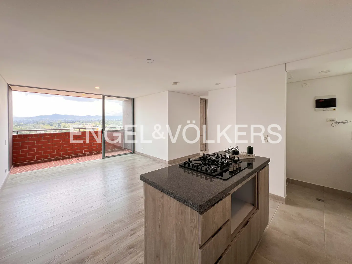 A modern, open-concept apartment with a kitchen island and a balcony with a brick railing overlooking a green landscape.