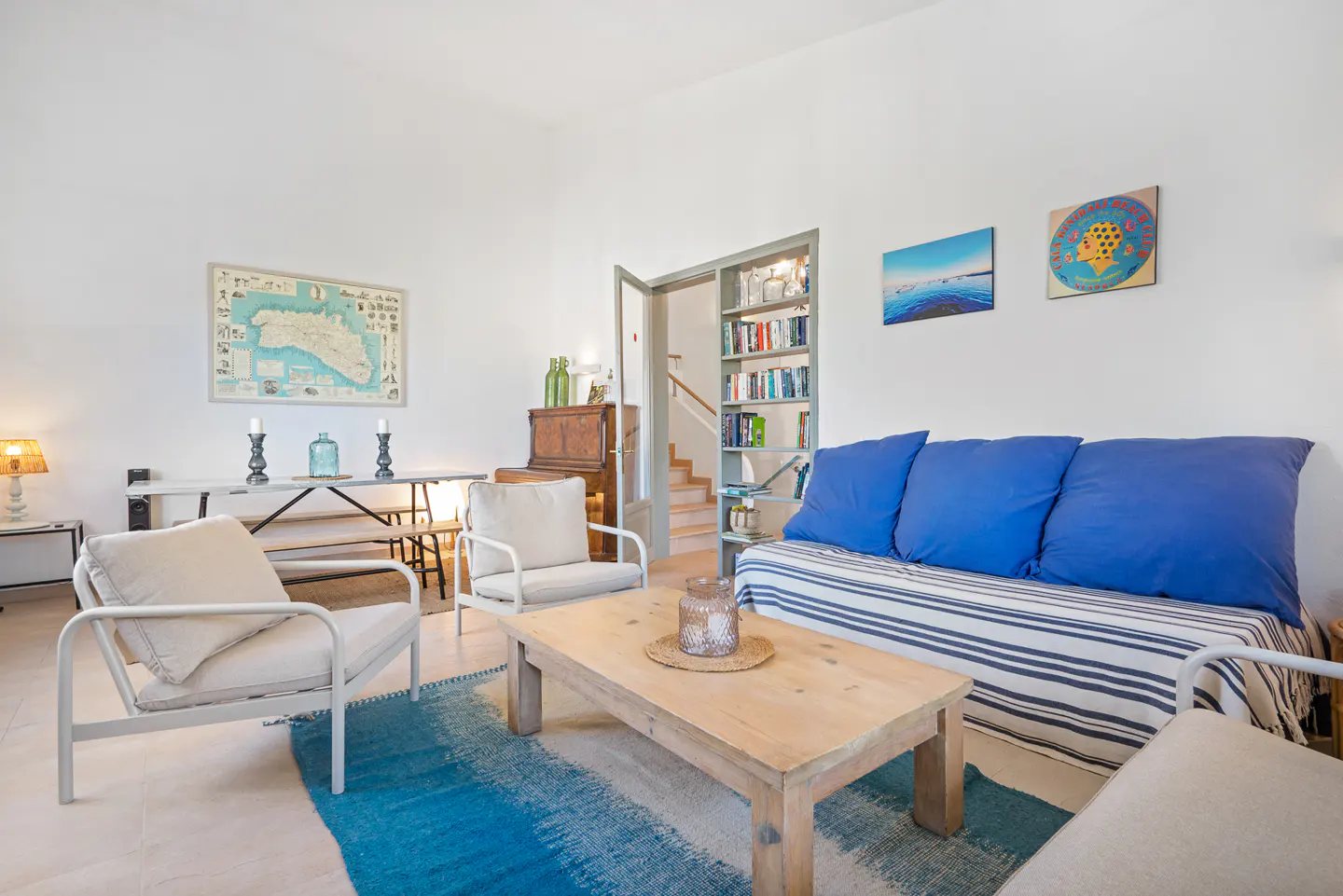 Bright living room with white walls, blue accents, and a wooden coffee table on a blue rug. A bookshelf and stairs are visible in the background.