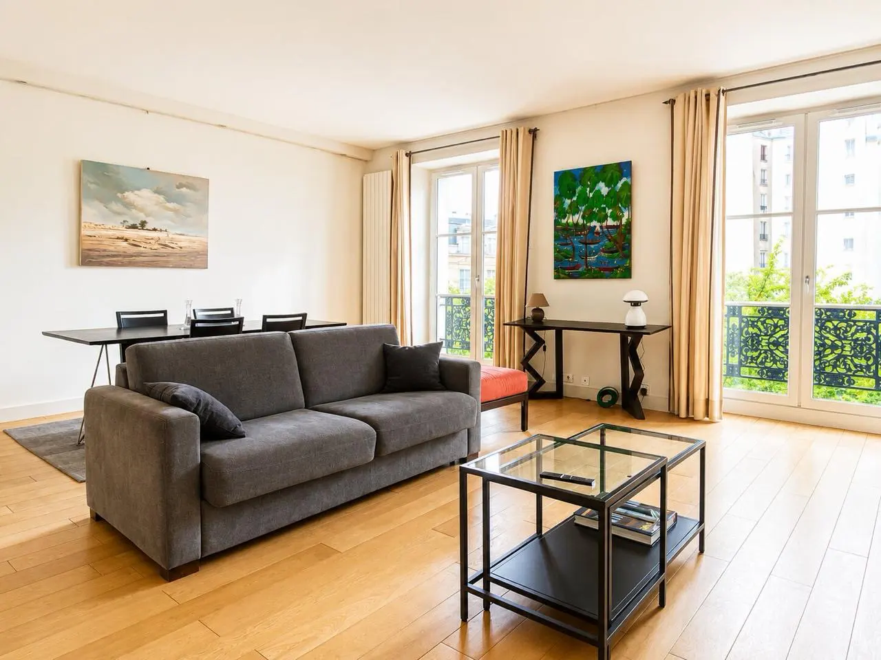 Bright living room with a grey sofa, glass coffee table, dining area, and large windows with beige curtains.