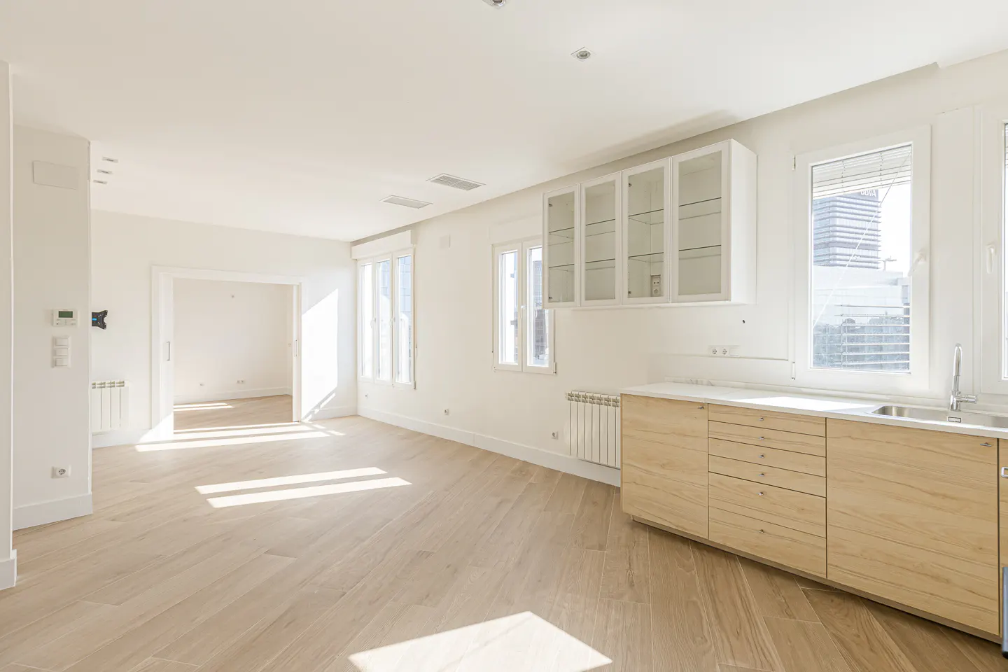 Bright, empty apartment with light wood floors, white walls, and kitchen cabinets. Sunlight streams through windows.