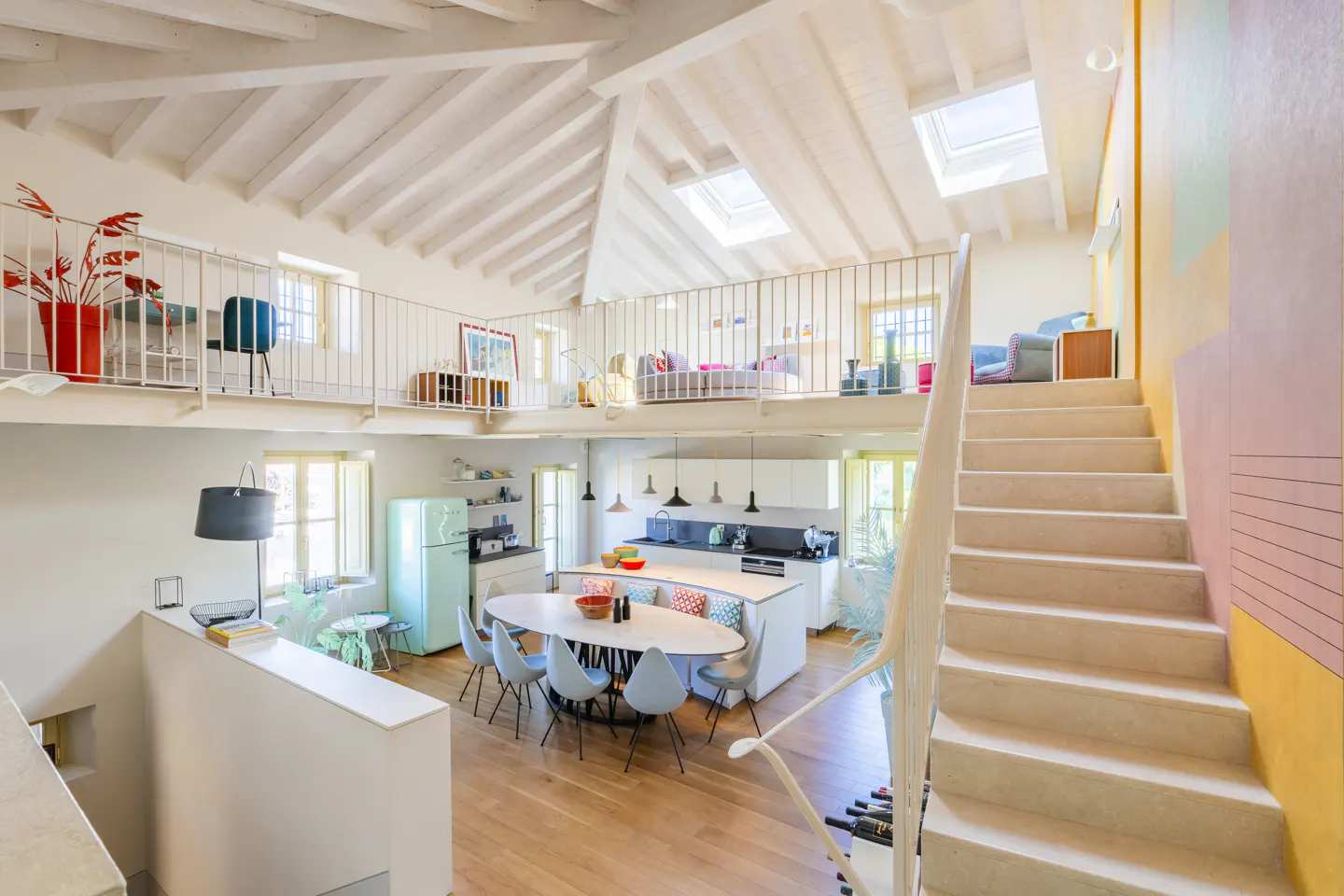 Bright, open-concept home interior with a loft, white walls, wood floors, and a modern kitchen with a light blue fridge. Stairs lead to the loft.