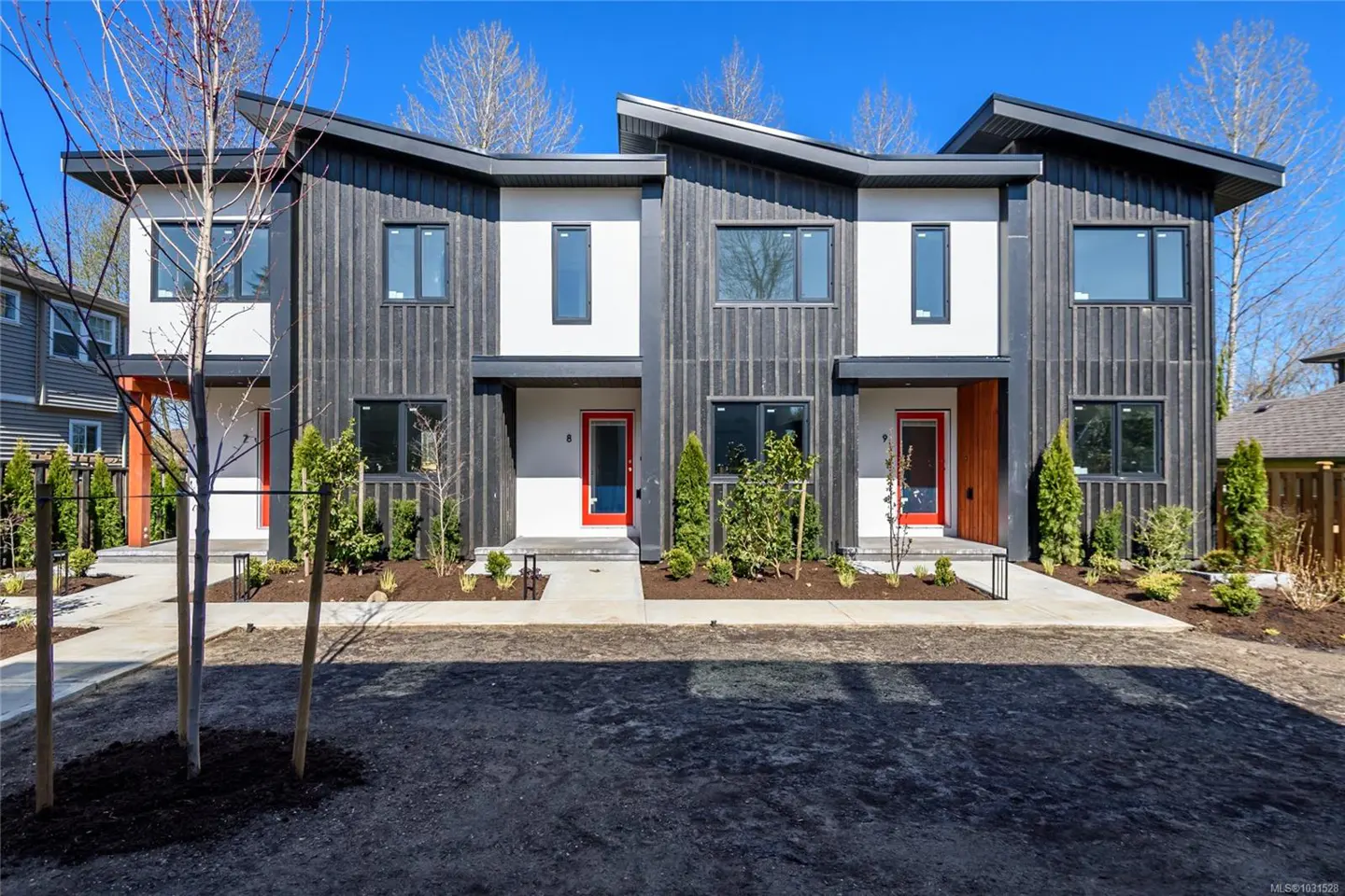 Modern townhouses with black siding, white accents, and red doors under a blue sky. Landscaped front yards with young trees.