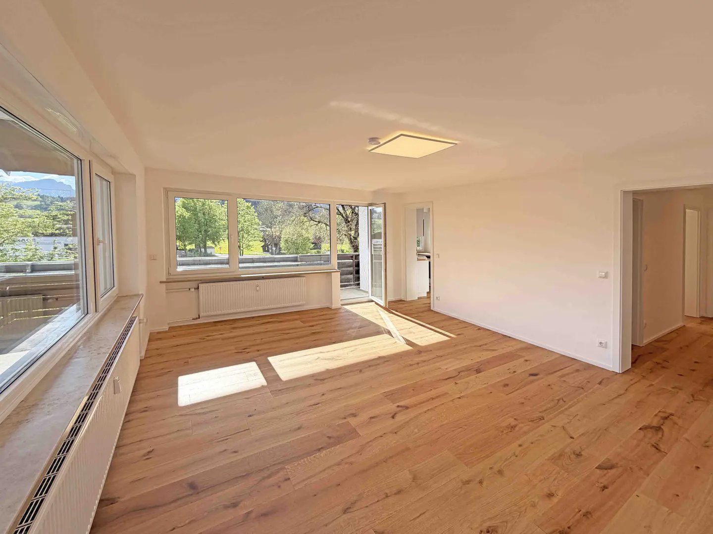 Bright, empty room with wood floors, white walls, and large windows showing trees and mountains. A door leads to a balcony.